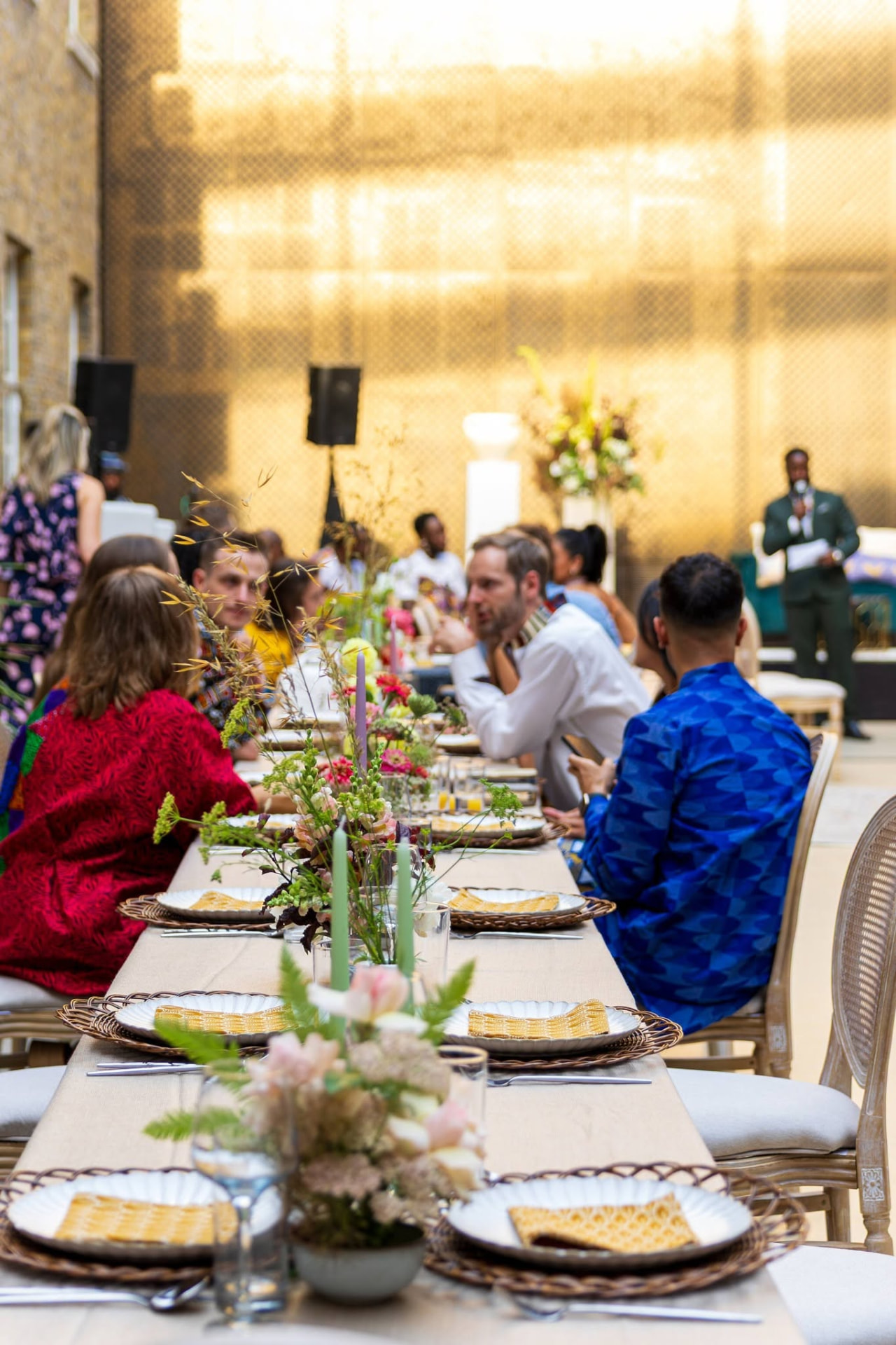 A long-table luncheon in a light-filled courtyard, set with woven chargers, garden florals and guests in conversation