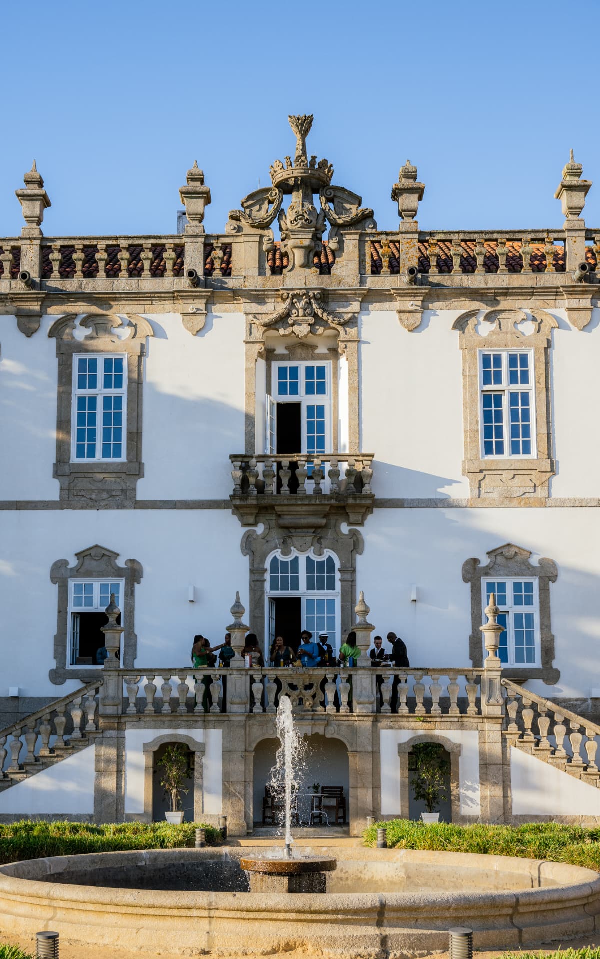 Guests gathering on a baroque palace balcony above a fountain at sunset